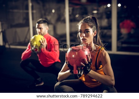 Close up of focused and motivated strong young fitness couple in sportswear crouching with the kettlebells reversed and doing squats in the gym.
