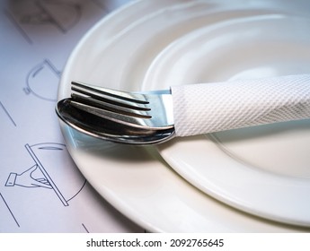 Close Up Of A Fork And Spoon Wrapped Neatly In A Paper Napkin And Placed On A White Ceramic Plate. 