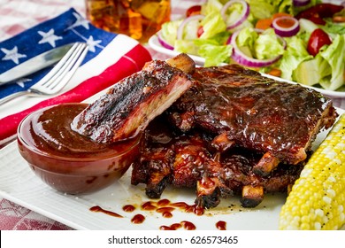 Close Up Of Fourth Of July Picnic With Barbeque Ribs Sitting On White Plate With Corn On The Cob, Salad And Glass Of Iced Tea