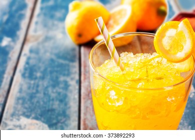 Close Up Of Fresh Orange Frozen Granita Slush Drink Garnished With Orange Wedge And Served In Glass With Striped Straw On Weathered Blue Wooden Picnic Table With Oranges In Background - Copy Space