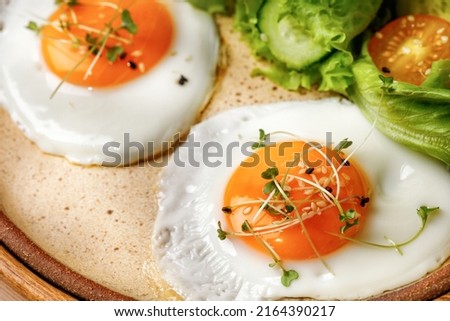 Close up of fried eggs with micro greens, breakfast with eggs, lettuce and cherry tomatoes. Beige ceramic plate.