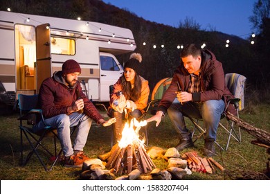 Close friends drinking beer together in the mountains and warming their hands around camp fire. Retro camper van with light bulbs. - Powered by Shutterstock