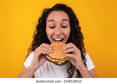 Close Up Front Portrait Of Hungry Female Model Eating Burger. Casual Young Lady Holding Snack Biting Tasty Fast Food Isolated On Orange Yellow Studio Wall, Banner. Guilty Pleasure Concept