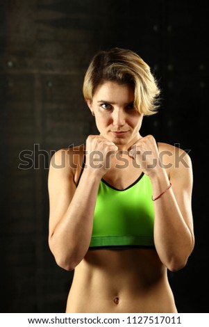 Close up front portrait of one young athletic woman in sportswear in gym over dark background, standing in boxing stance with hands and fists, looking at camera