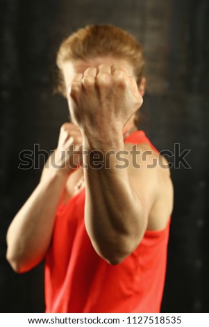 Close up front portrait of one young middle age athletic woman in sportswear in gym over dark background, standing in boxing stance with hands and fists, looking at camera