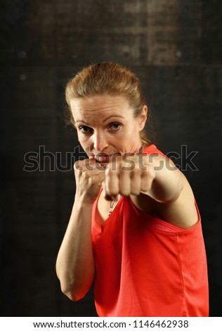 Close up front portrait of one young mid adult athletic woman in sportswear in gym over dark background, standing in boxing stance with hands and fists, looking at camera