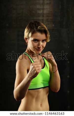 Close up front portrait of one young athletic woman in sportswear in gym over dark background, standing in boxing stance with hands and fists, looking at camera