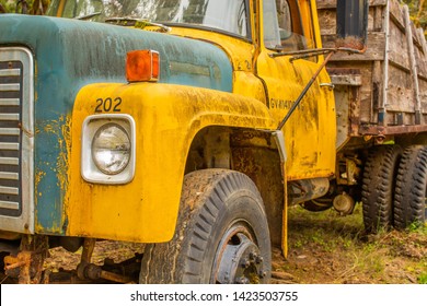 A Close Up Of The Front Of A Rusted Yellow And Blue Vintage Industrial Truck With A Wooden Flat Bed Sitting Abandoned In An Old Work Yard.