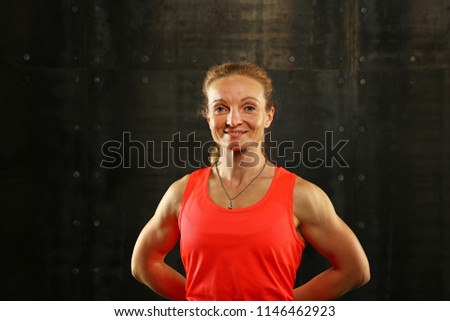 Close up front upper body portrait of one middle age athletic woman in sportswear in gym over dark background, looking at camera and smiling