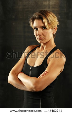 Close up front upper body portrait of one young athletic woman in sportswear in gym over dark background, looking at camera