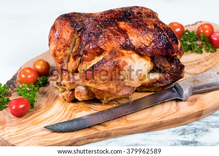 Close up front view of barbecued chicken with vegetables, herbs and large cutting knife on wooden server with white marble underneath. Selective focus on upper front leg of chicken. 