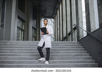 Close Up Front View Of Pleasant Young Asian Indian Doctor In White Coat, Holding A Clipboard, Standing On Stairs Of Modern Clinic Outdoors, Smiling And Looking At Camera. Full Length Shot