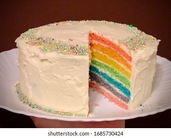 Close Up Full Frame Photograph Of A 7 Layer Rainbow Cake With Sprinkles And White Buttercream With Dark Background On A White Plate