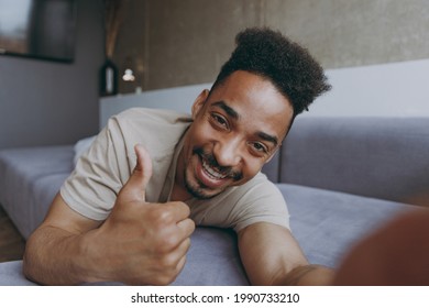 Close Up Full Length Young African American Man In Casual Beige T-shirt Sweatpants Lay Down On Grey Sofa Show Thumb Up Indoors Partment Do Selfie Shot On Mobile Phone Rest On Weekends Staying At Home