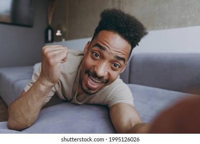 Close Up Full Length Young African American Man In Casual Beige T-shirt Sweatpants Lay Down On Grey Sofa Do Winner Gesture Indoors Partment Do Selfie Shot On Mobile Phone Rest On Weekends Stay At Home