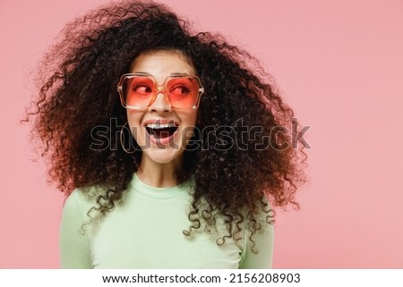Close up fun joyful young curly latin woman 20s years old wears mint t-shirt sunglasses looking aside isolated on plain pastel light pink background studio portrait. People emotions lifestyle concept