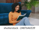 Close up fun little kid teen girl wear casual clothes reading novel paper book sits near blue sofa couch stay at home hotel flat rest relax spend free spare time in living room indoor. Lounge concept