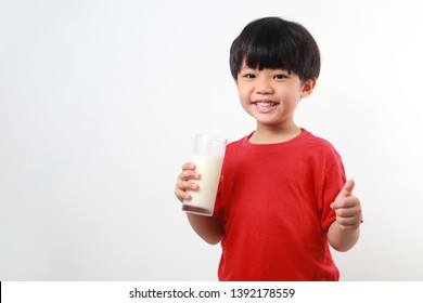 Close Up Fun Portrait Of Cute Little Asian Boy Drinking Milk And Showing Thumb Up Sign. Isolated Against Light Background.