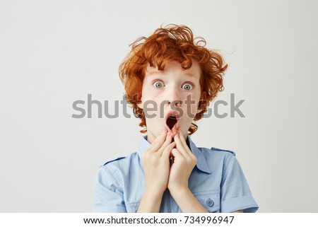 Close up of funny by with curly ginger hair and freckles holding hands near mouth, being shocked seeing adult people kissing.