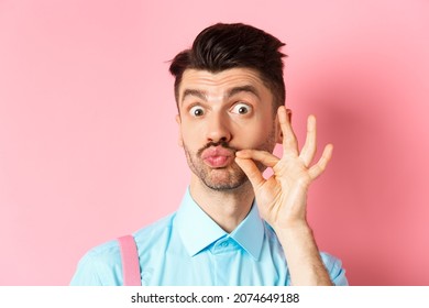 Close up of funny caucasian guy touching his french moustache, pucker lips and looking silly, standing over pink background