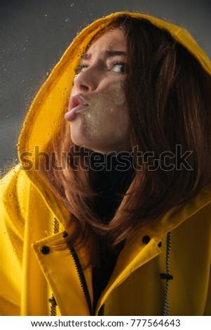 Close up of a funny girl dressed in raincoat with a hood on her head standing behind a wet with rain window and looking at camera isolated over gray background