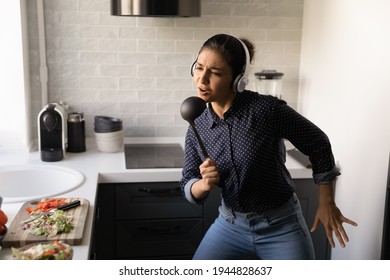 Close Up Funny Indian Woman Wearing Headphones Singing Into Kitchenware As Microphone, Dancing To Favorite Music In Modern Kitchen At Home, Overjoyed Young Female Having Fun, Preparing Meal