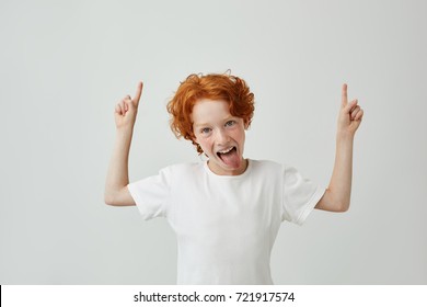 Close up of funny little boy with curly ginger hair and freckles pointing up with both hands, having silly face with open mouth. Copy space.