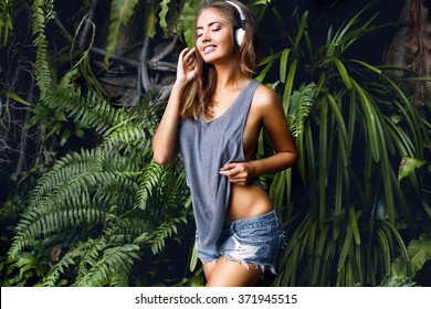 Close up funny portrait of young pretty hipster blonde girl listening music on big white earphones,wearing mini denim t-shirt,posing at city center at sunny summer, smiling,teen,cool accessories,laugh