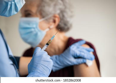 Close Up Of General Practitioner Hand Holding Vaccine Injection While Wearing Face Protective Mask During Covid-19 Pandemic. Young Woman Nurse With Surgical Mask Giving Injection To Senior Woman.