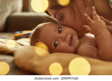 Close Up Gentle Kiss Of Affectionate Mother. Smiling Baby Looking At Camera, Feeling Mother's Kiss On Its Neck. Side View Woman And Child Faces, Warm Lights With Bokeh Effect.