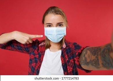 Close Up Girl In Checkered Shirt Isolated On Red Background. Epidemic Pandemic Coronavirus 2019-ncov Sars Covid-19 Flu Virus Concept. Doing Selfie Shot On Mobile Phone Point Index Finger On Face Mask