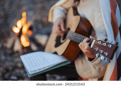 Close up of a girl guitarist practicing on nature near the camp fire. Warm evening shot of a girl playing the guitar with notebook, composing new song near bonfire - Powered by Shutterstock