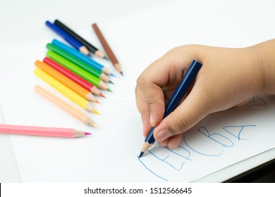 Close Up Of Girl Hand With Pencil Writing English Words By Hand On  White Notepad Paper At White Background, A Child Is Holding A Crayon In His Hands At Paper And Crayons For Drawing
