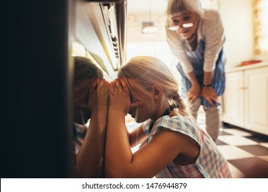 Close Up Of A Girl Looking Inside An Oven Through The Glass Door. Granny And Kid In Kitchen Looking For Cake Inside The Oven.