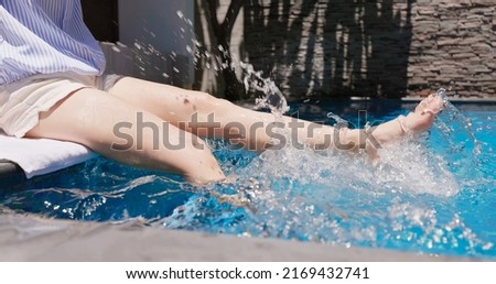 close up of girl is sitting on edge of swimming pool kicking feet underwater in rippled clear blue water 