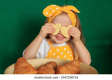 Close up glad joyful laughing child closing eyes by two cookies pastry in shape of heart in her hands, indulges. Little girl in yellow apron posing on green background. Concept Easter holliday baking
