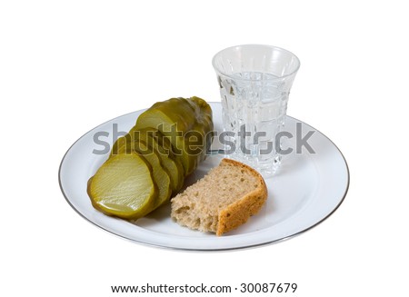 A close up of the glass of vodka, pickled cucumber and piece of black bread.