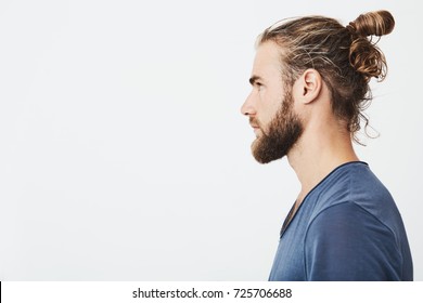 Close up of good-looking bearded hipster guy with hair in bun, in blue t-shirt standing in profile, looking aside, posing for photo. - Shutterstock ID 725706688