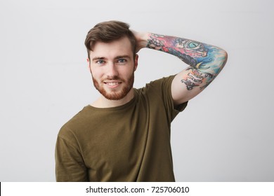 Close Up Of Good-looking Cool European Guy With Beard And Arm Tattoo Smiling Brightfully, Holding Hand Behind Head With Cheerful Expression, Posing For University Graduation Photo Album.
