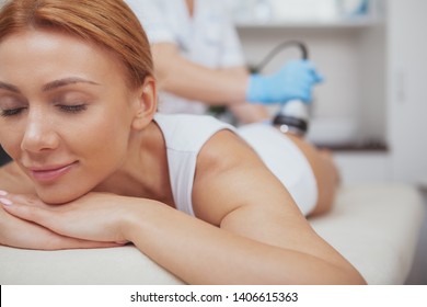Close up of a gorgeous happy woman smiling, receiving ultrasound cavitation treatment by cosmetologist, copy space. Charming woman enjoying body contouring therapy at spa clinic