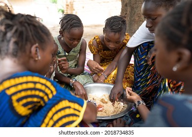 Close up of a goup of coulorfully dressed black girls sitting around a big metal bowl, sharing a typical African meal made out of rice and vegetables - Powered by Shutterstock
