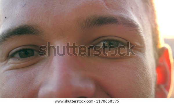 Close up of green eyes of an young happy guy\
blinking and looking into camera against the background of cars\
parking. Portrait of handsome man standing in urban environment and\
smiling at sunset.