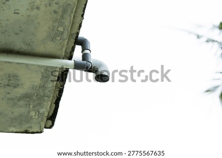 Close Up Of A Grey Gutter Pipe With White Downspout Attached To A Dirty Concrete Eaves Against A Bright White Sky With Green Palm Leaves In The Upper Right Corner