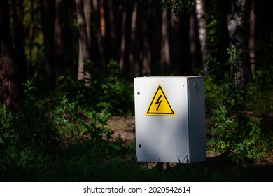 Close Up Grey Hinged Power Supply Box In The Forest. Outside Electrical Box With Yellow Danger Warning Sign. Electricity In Remote Places.