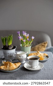 Close up of a grey tablecloth setting with two plates of croissants, apricot jam, a vintage porcelain cup of coffee, and small pots of spring flowers in soft light.