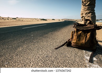 Close up ground point of view of man with leather backpack travel and wait for a car to share the trip together - sand and desert dunes and black asphalt road for traveler people enjoying lifestyle