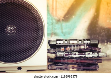 Close Up Of A Group Of Cassettes Stacked Next To The Speaker Of An Old Boom Box And On A Graffiti Background. Listening To The Music Of The Past.