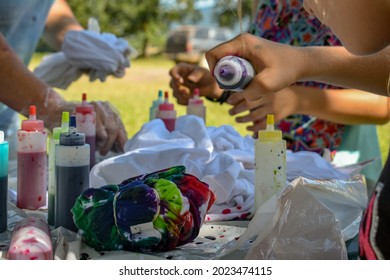 A Close Up Of A Group Of People Making Tie Dye Tee Shirts Outside On A Table- A Fun Day Outside Making Tie Dye Shirts- Colorful Bottles Of Dye On A Table Outside- A Group Of Kids Doing Artwork