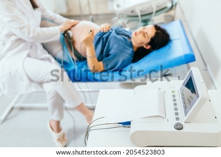 Close up of gynecologist doing electrocardiograph check up for pregnant woman at clinic. Focus on modern equipment with charts of baby's heartbeat. Blur background.