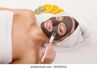 Close up of hand of beautician applying chocolate mask on female face with brush. The pretty young woman is lying and smiling. She closed eyes with pleasure